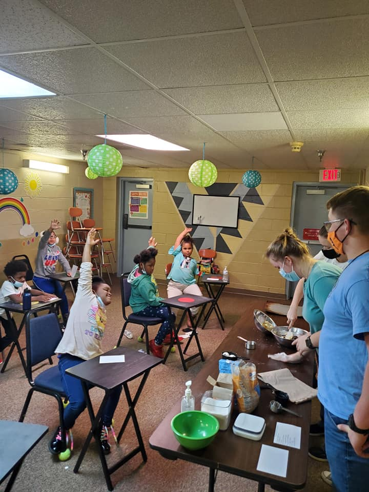 students in a classroom raising their hands and teachers at the front of the room doing a science experiment