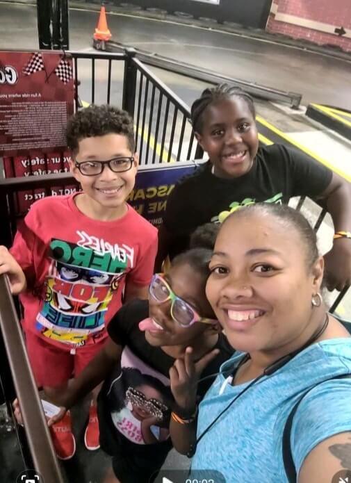 A lady smiling with three young children while waiting in a line to race go-karts.