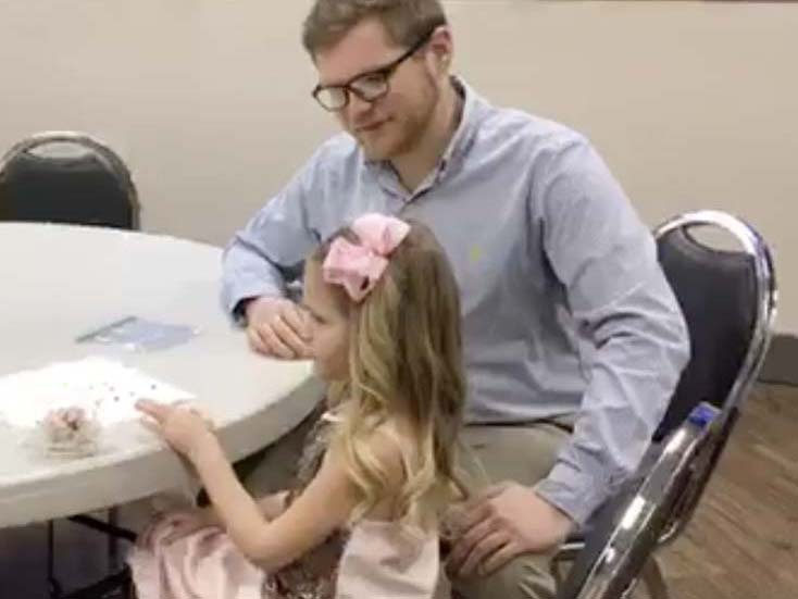 A father sits with his daughter and is watching her talk and do crafts.