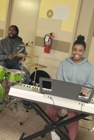 A woman sits behind a piano while a man plays the drums.