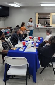 A group of people at a table listen to a woman talking.