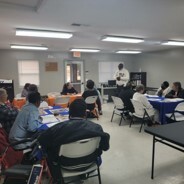 A large group of people in a room sitting at various tables and listening to a speaker. 